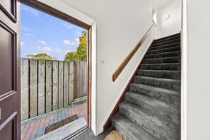 Una escalera con alfombra gris que sube a un piso superior, una puerta de madera, y vistas a una valla de madera y vegetación al exterior.