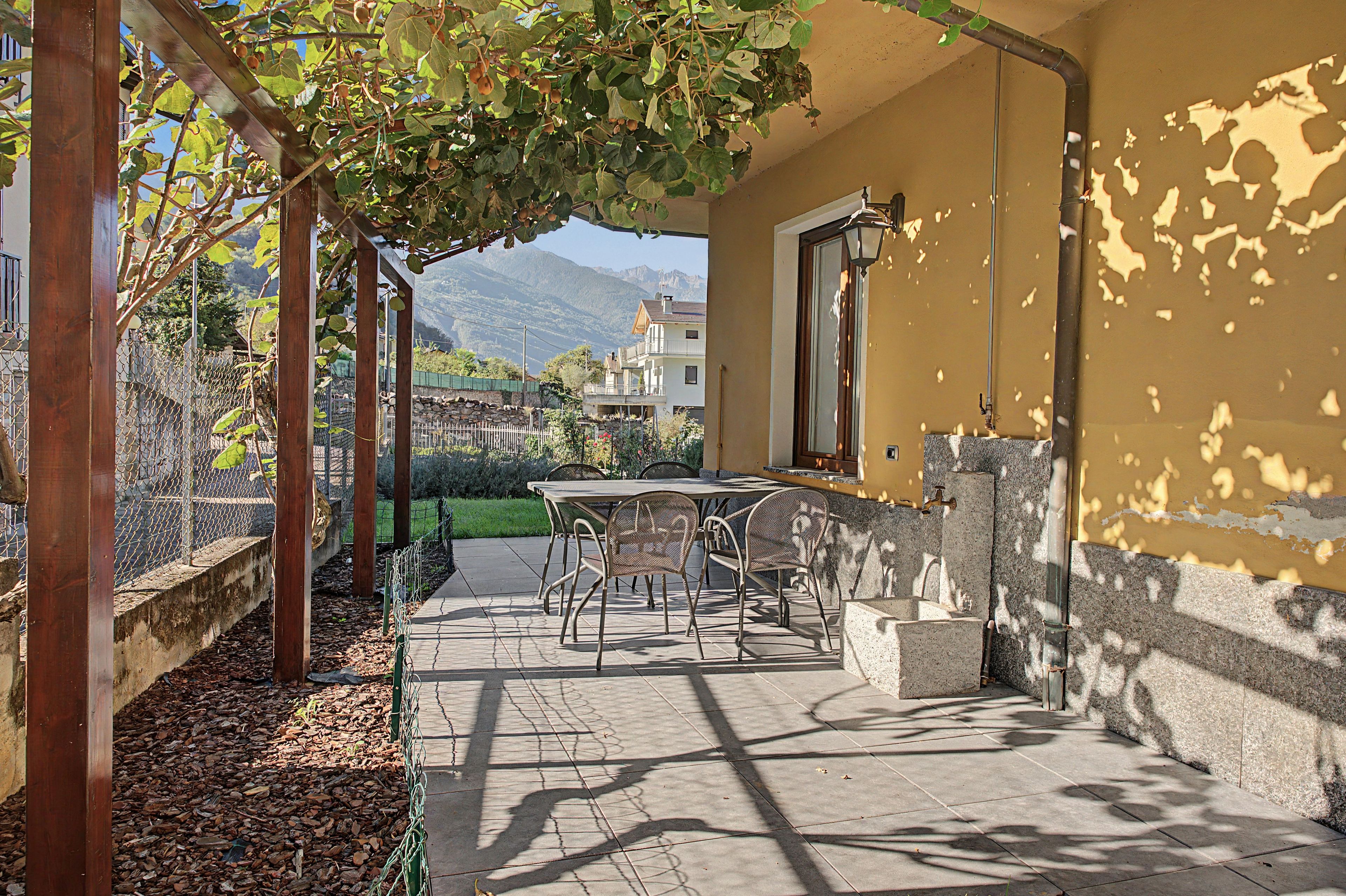 Un patio con tavolo e sedie, una pergola ricoperta di piante rampicanti e vista su una montagna sullo sfondo.