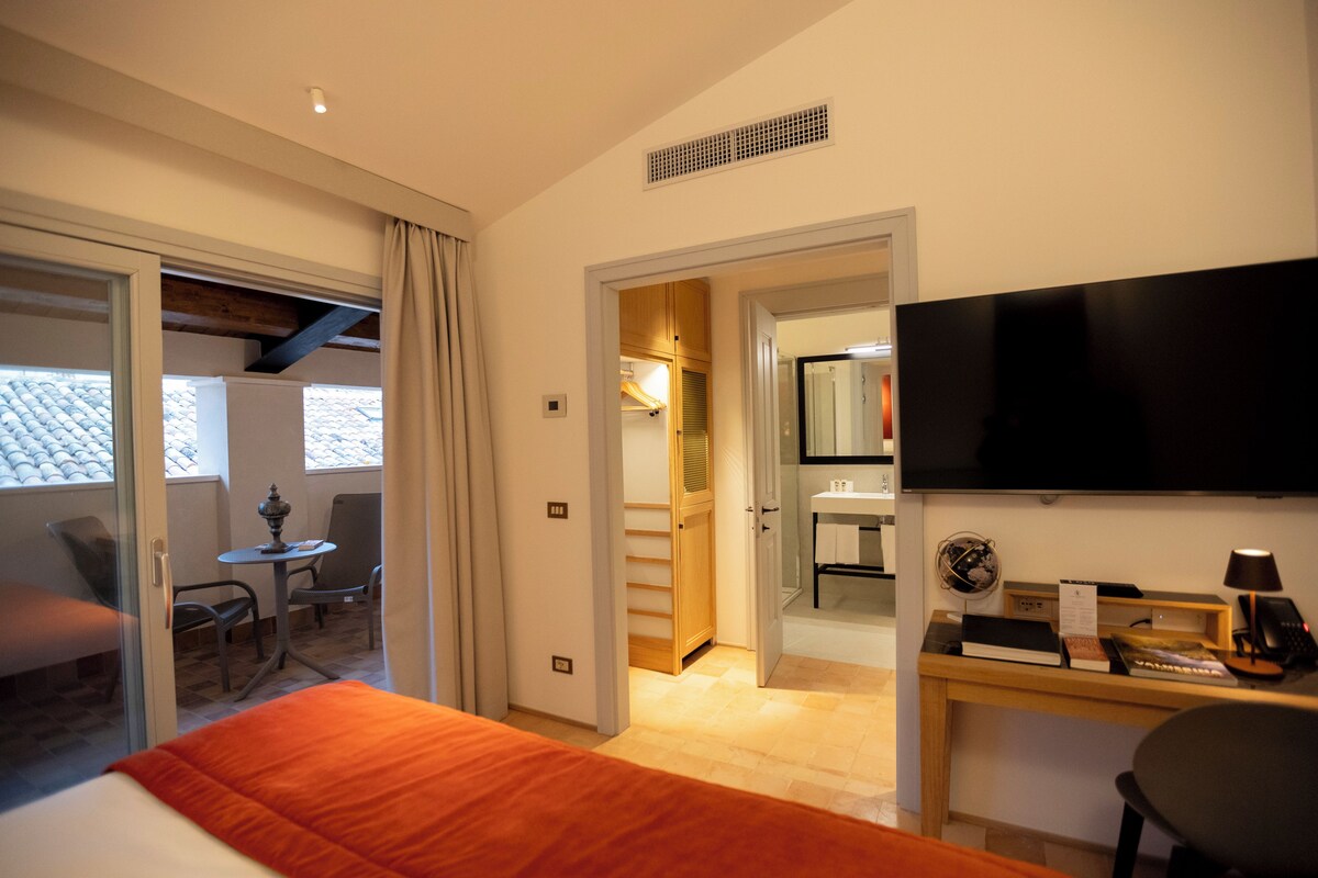 A hotel room with a bed, a desk with a flat-screen TV, a chair, and a small table.