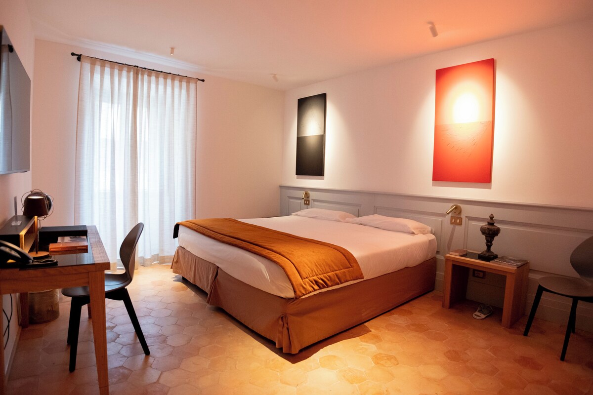 A hotel room with a bed, a desk, a chair, a nightstand, and a wall-mounted lamp.