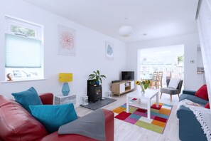 Ein modernes Wohnzimmer mit einer Couch, einem Couchtisch und einem Fernseher. Ein Fenster mit Jalousien, eine Lampe und eine Pflanze sind ebenfalls vorhanden.