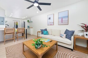 A modern living room with a sofa, coffee table, and chairs. There is a ceiling fan, a kitchen area in the background, and a beach-themed wall art. - Kahaluu Beach Surf, Oceanview, 2bed,2bath, Lanai (Kailua-Kona)