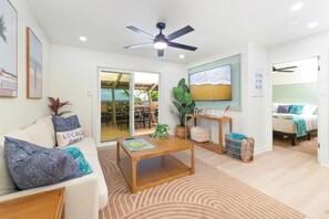 Uma sala de estar moderna com sofá, mesa de centro e ventilador de teto. Há uma porta que dá para uma área externa com mesa e cadeiras.
