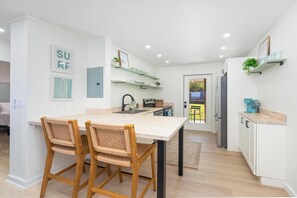Cozinha moderna com armários brancos, área de refeições com duas cadeiras de vime e vista pela porta.