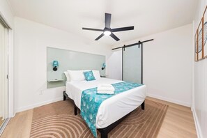 Quarto de hotel com cama, ventoinha no teto, mesa de cabeceira, espelho e janela.