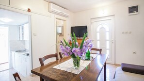 A dining area with a wooden table, chairs, and a vase of purple flowers. There is a kitchen visible in the background with a refrigerator and a television. - House with large garden, right next to the river (Žrnovnica)