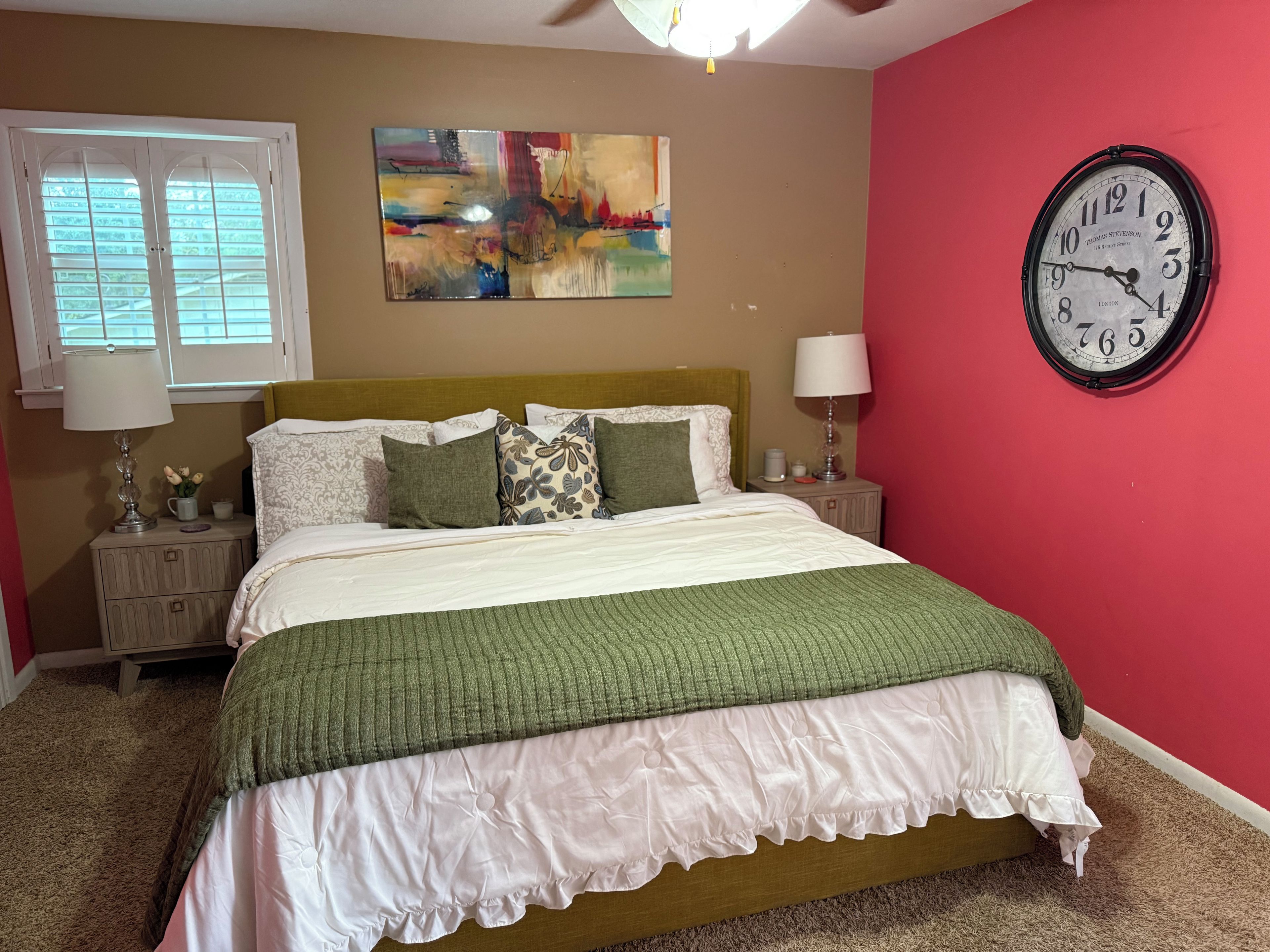 Une chambre à coucher avec un lit, une horloge murale, un tableau et une fenêtre avec des stores.
