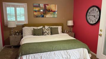A bedroom with a bed, two bedside tables, a wall clock, a window with blinds, and a painting.