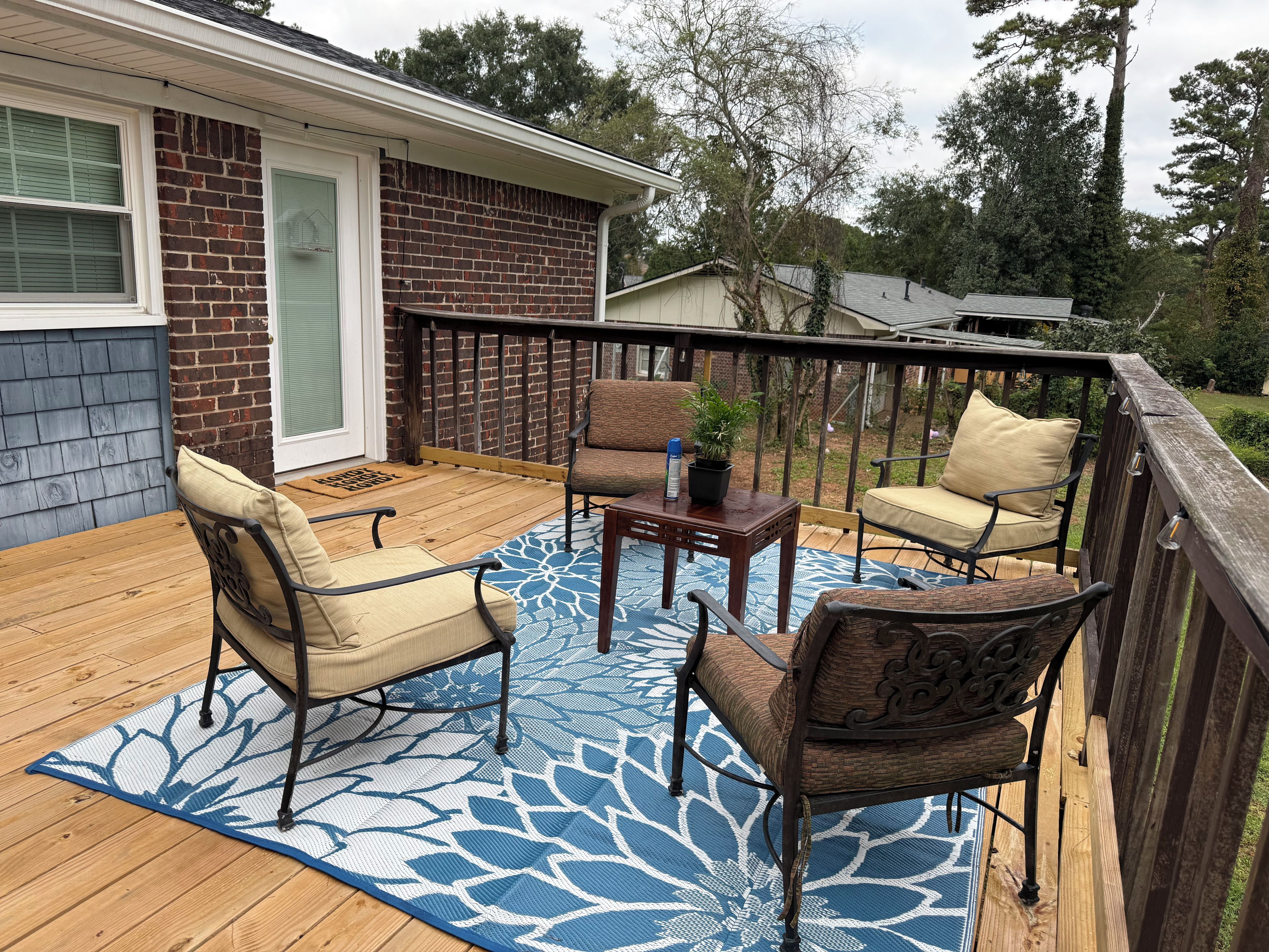 An outdoor deck with a blue patterned rug, two chairs, a small table, and a potted plant.