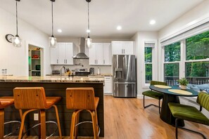 A modern kitchen with a central island, stainless steel appliances, and a dining area with green upholstered chairs. - Arden Legacy Retreat: Pool, Sauna, Hot Tub, Game Room, Theater (Avery Creek)