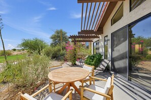 Eine Terrasse im Freien mit einem runden Holztisch und Stühlen, umgeben von Pflanzen und einer Pergola.