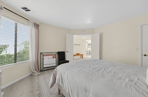 Quarto com cama, cadeira, mesa de cabeceira, janela com persianas e vista para o exterior.