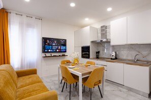 Ein modernes Wohnzimmer mit einer Couch und einem Esstisch, eine Küche mit weißen Schränken und einem an der Wand montierten Fernseher.