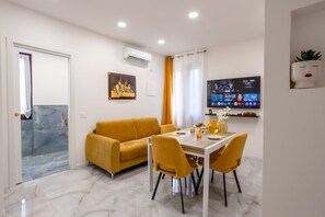 Ein modernes Wohnzimmer mit einer Couch, einem Esstisch und Stühlen. Ein Flachbildfernseher ist an der Wand montiert, darüber befindet sich eine Klimaanlage.