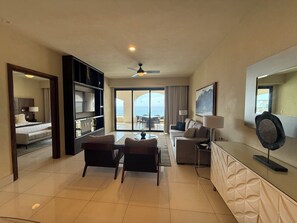 Habitación de hotel con balcón, zona de estar con sillas y mesa, sofá, televisión y ventilador de techo.