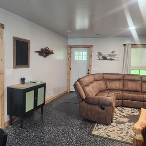 Ein Wohnzimmer mit einem Ecksofa, einem Holzkabinett, einem Wandbild und einer Tür mit Fenster.