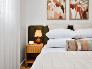 A neatly made bed with white bedding, a bedside lamp, and a small wooden nightstand with a bowl and a lamp. There are two paintings on the wall above the bed. - Olive & Ochre | Warm Coastal Retreat (Shoalwater)