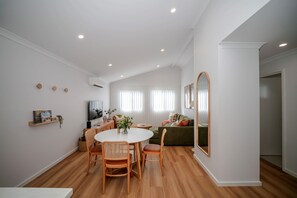 A modern living room with a dining area, a sofa, a TV, and a large mirror. - Olive & Ochre | Warm Coastal Retreat (Shoalwater)