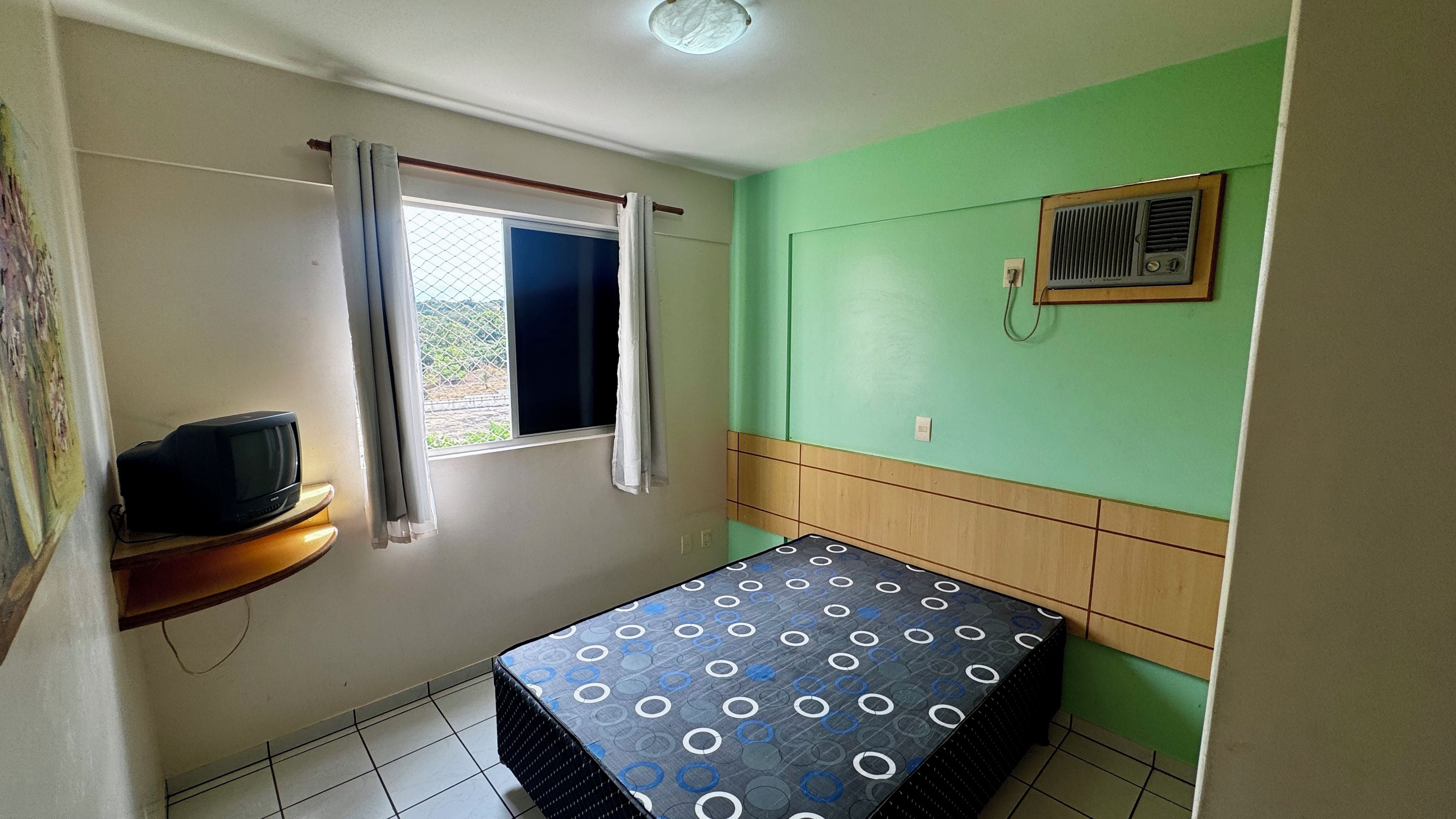 Een kleine, eenvoudige kamer met een bed, een raam met gordijnen, een wandheater en een televisie op een houten dressoir.