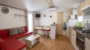 A compact living space with a red sofa, a small dining table, a kitchenette, and a wall-mounted TV. - Mobile Home 'Location' with Shared Pool, Wi-Fi and Air Conditioning (Argelès-sur-Mer)
