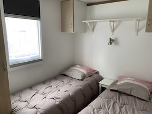 A small, single-room accommodation with two beds, a window, a white shelf, and a door. - Mobile Home 'Location' with Shared Pool, Wi-Fi and Air Conditioning (Argelès-sur-Mer)