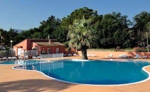 A swimming pool with lounge chairs, a palm tree, and a building with a red facade. - Mobile Home 'Location' with Shared Pool, Wi-Fi and Air Conditioning (Argelès-sur-Mer)