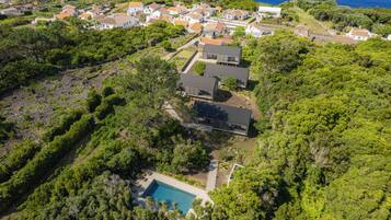 A residential area with a large house featuring a swimming pool, surrounded by dense greenery and a coastal view.
