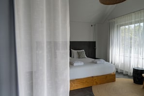 A modern bedroom with a large bed, a headboard, and a view of the outdoors through sheer curtains.