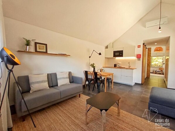 Un salon moderne avec un canapé, une petite table et un coin repas avec des chaises. Au fond, on aperçoit une cuisine équipée d’un micro-ondes et de placards.