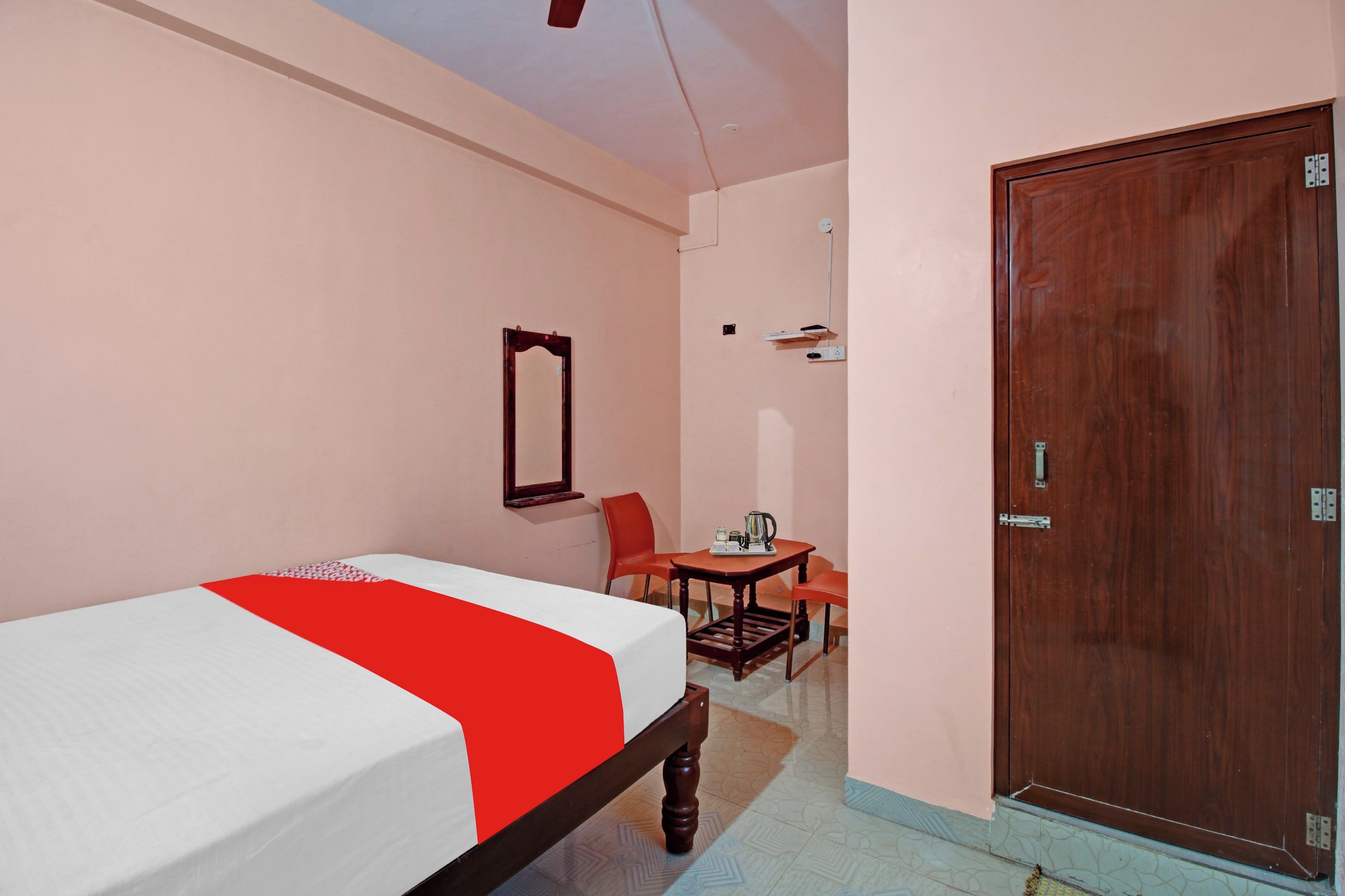 A bedroom with a bed, a wooden door, a small table with a chair, and a mirror on the wall.