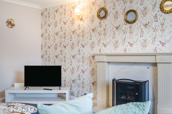A living room with a television on a stand, a fireplace, and a patterned wallpaper.