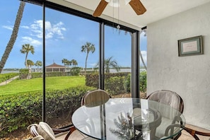 A room with a glass table and chairs, large windows with a view of a lawn and palm trees, and a ceiling fan. - Sunrise Cove 103: Ground Floor Waterfront Condo on Siesta Key w/ Sleeper Sofa (Sarasota)