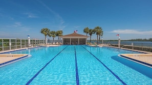 Une piscine à l’eau d’un bleu cristallin, entourée de chaises longues et de palmiers, avec un espace de repos couvert situé au centre.