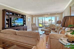 A living room with a large flat-screen TV, beige sofas, a glass coffee table, and a view of the outdoors. - Sunrise Cove 107: Ground Floor Waterfront Condo on Siesta Key w/ Sleeper Sofa (Sarasota)