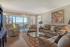 A spacious living room with a glass coffee table, a sofa, and a recliner. There is a dining area with a table and chairs, and a large window offering a view of the outdoors. - Sunrise Cove 107: Ground Floor Waterfront Condo on Siesta Key w/ Sleeper Sofa (Sarasota)