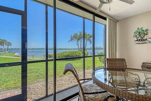 Quarto com mesa e cadeiras de vidro, vista para um corpo d'água e ventilador de teto.