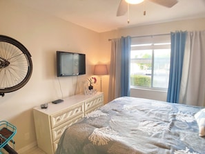 A hotel room with a bed, a television mounted on the wall, a bedside table with a lamp, and a window with curtains. - Sunrise Cove 126: Remodeled Waterfront Condo on Siesta Key w/ Sleeper Sofa (Sarasota)