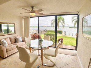 A room with a glass table, two chairs, a sofa, a ceiling fan, and a large window with a view of a body of water and greenery. - Sunrise Cove 126: Remodeled Waterfront Condo on Siesta Key w/ Sleeper Sofa (Sarasota)