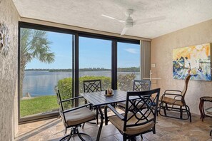 Ein Essbereich mit Tisch und Stühlen, große Fenster mit Blick auf ein Gewässer und viel Grün sowie ein Deckenventilator.