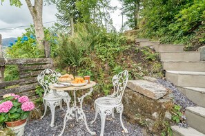 A rustic outdoor seating area with a round table and two chairs, surrounded by greenery and a stone wall. - Two Bedroom Holiday Cottage At The Foot Of Yr Wyddfa (Snowdon) | Clogwyn-Bach (Rhyd Ddu)