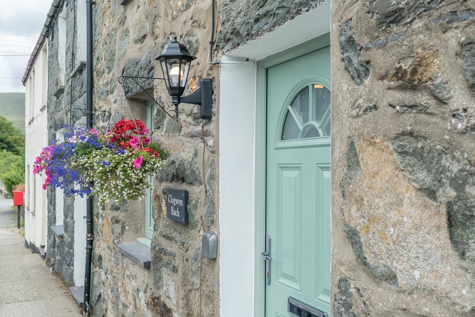 A stone building with a green door, a hanging flower basket, and a black wall-mounted light fixture.