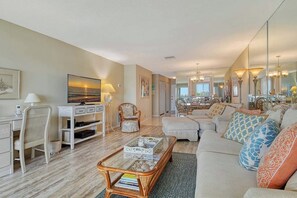 A living room with a sofa, coffee table, and chairs, a dining area with a table and chairs, and a large mirror reflecting the room. - Sunrise Cove 201: Waterfront Condo on Siesta Key with Heated Lap Pool (Sarasota)