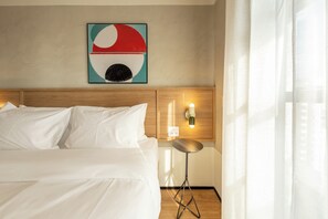 A hotel room with a bed, a wall-mounted artwork, a bedside table with a lamp, and a curtain. - Librio by Bhomy (São Paulo)