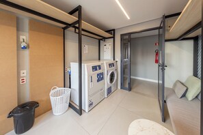 A laundry area with a washing machine, a drying machine, a trash bin, and a basket. - Librio by Bhomy (São Paulo)