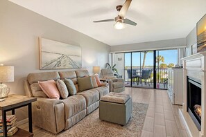 A living room with a sofa, ottoman, and a fireplace. There is a ceiling fan and a painting on the wall. - Sunrise Cove 203: Remodeled Waterfront Condo on Siesta Key w/ Sleeper Sofa (Sarasota)