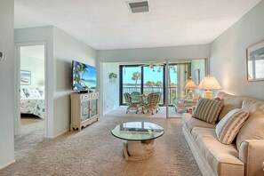 A living room with a glass coffee table, a beige sofa, a television, and a view of the outdoors through large windows. - Sunrise Cove 204: Waterfront Vacation Condo on Siesta Key with Sleeper Sofa (Sarasota)
