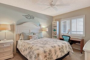 Ein Schlafzimmer mit Bett, Nachttischen, Schreibtisch, Stuhl, Deckenventilator und einem Fenster mit Jalousien.