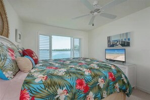 Een slaapkamer met een bed, een plafondventilator, een televisie en een raam met uitzicht op het strand.
