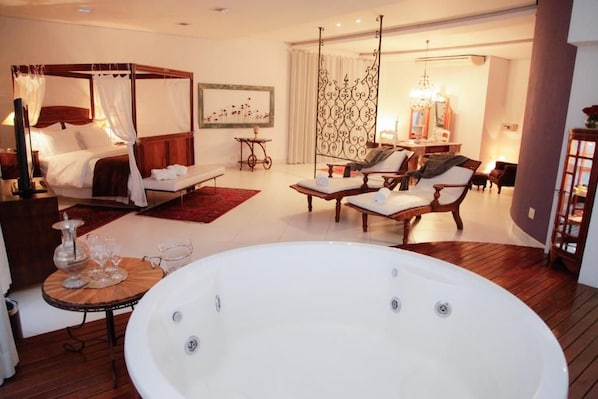 A bedroom with a four-poster bed, a sitting area with chairs, a large bathtub, and a dining area with a chandelier.