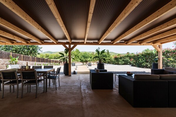Een terras buiten met een houten dak, een tafel met stoelen en een zithoek met kussens.
